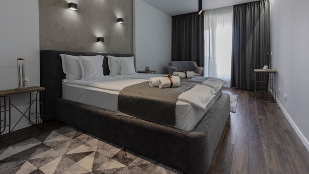 A modern hotel room with a large bed, a bedside table, a chair, and a small table with a lamp. - Ares Aparthotel (Cluj-Napoca)