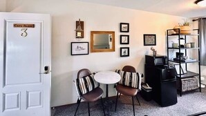 A compact living room with a white door, a small round table, two chairs, a black entertainment unit, and a wall-mounted mirror.