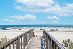 Ein hölzerner Steg führt zu einem Sandstrand mit Sonnenschirmen und Badegästen.