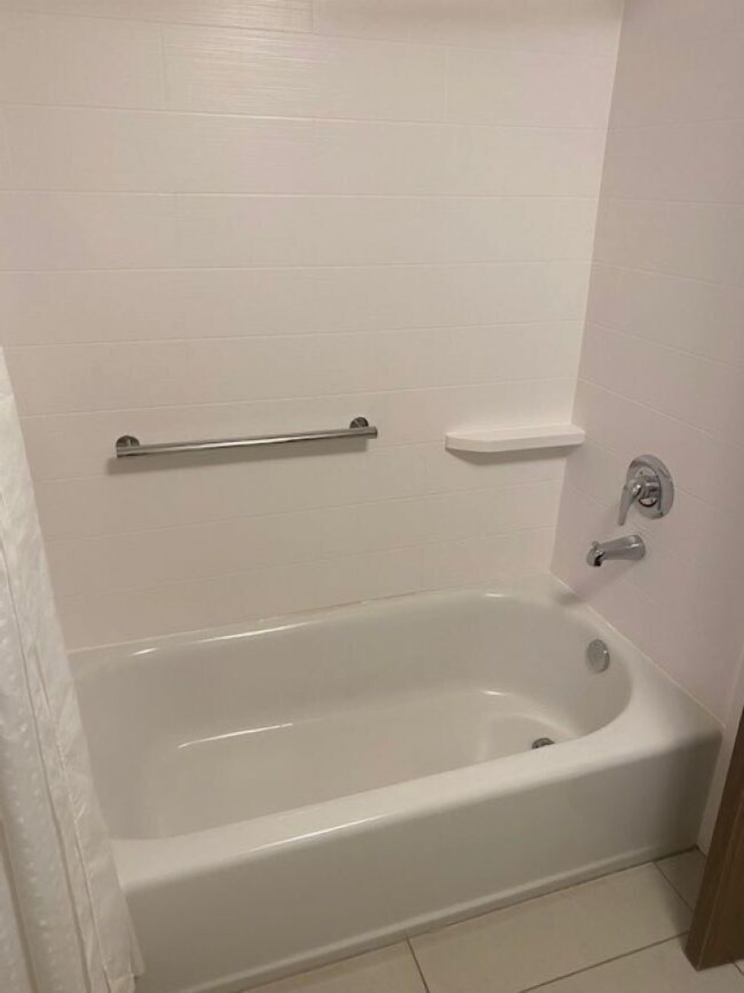 A standard white-tiled bathroom with a bathtub, a grab bar, and a small shelf.