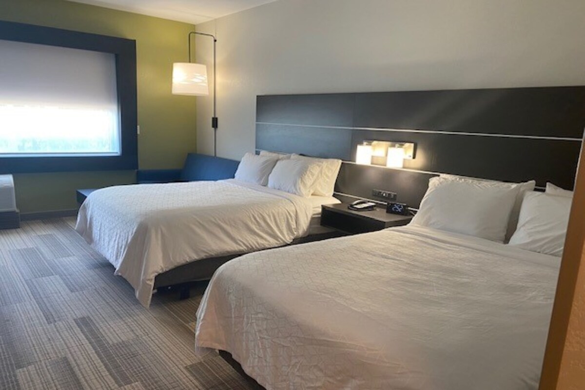 A hotel room with two beds, a nightstand, a lamp, and a window with blinds.