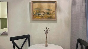 Ein Zimmer mit einem runden Tisch, zwei Stühlen, einer Vase mit Trockenpflanzen und einem gerahmten Gemälde an der Wand.