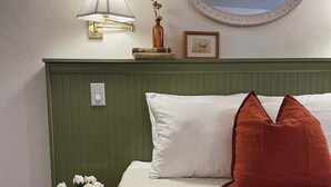 Ein ordentlich bezogenes Bett mit weißen und roten Kissen, ein Nachttisch mit Blumenvase, eine Wandlampe und ein gerahmtes Bild.