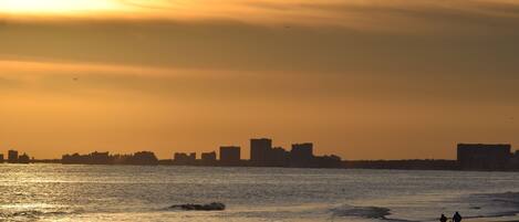 A beach with a sunset, waves, and silhouettes of buildings in the distance.