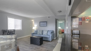 A living room with a blue sofa, a patterned armchair, and a coffee table. There is a kitchen area with a refrigerator and a toaster. - Waipani E13 - 2BR Charming Condo! Across from Beach in Ocean Drive! (North Myrtle Beach)
