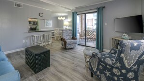 A living room with a sofa, a chair, a small table, and a kitchen area with a bar counter. - Waipani E13 - 2BR Charming Condo! Across from Beach in Ocean Drive! (North Myrtle Beach)