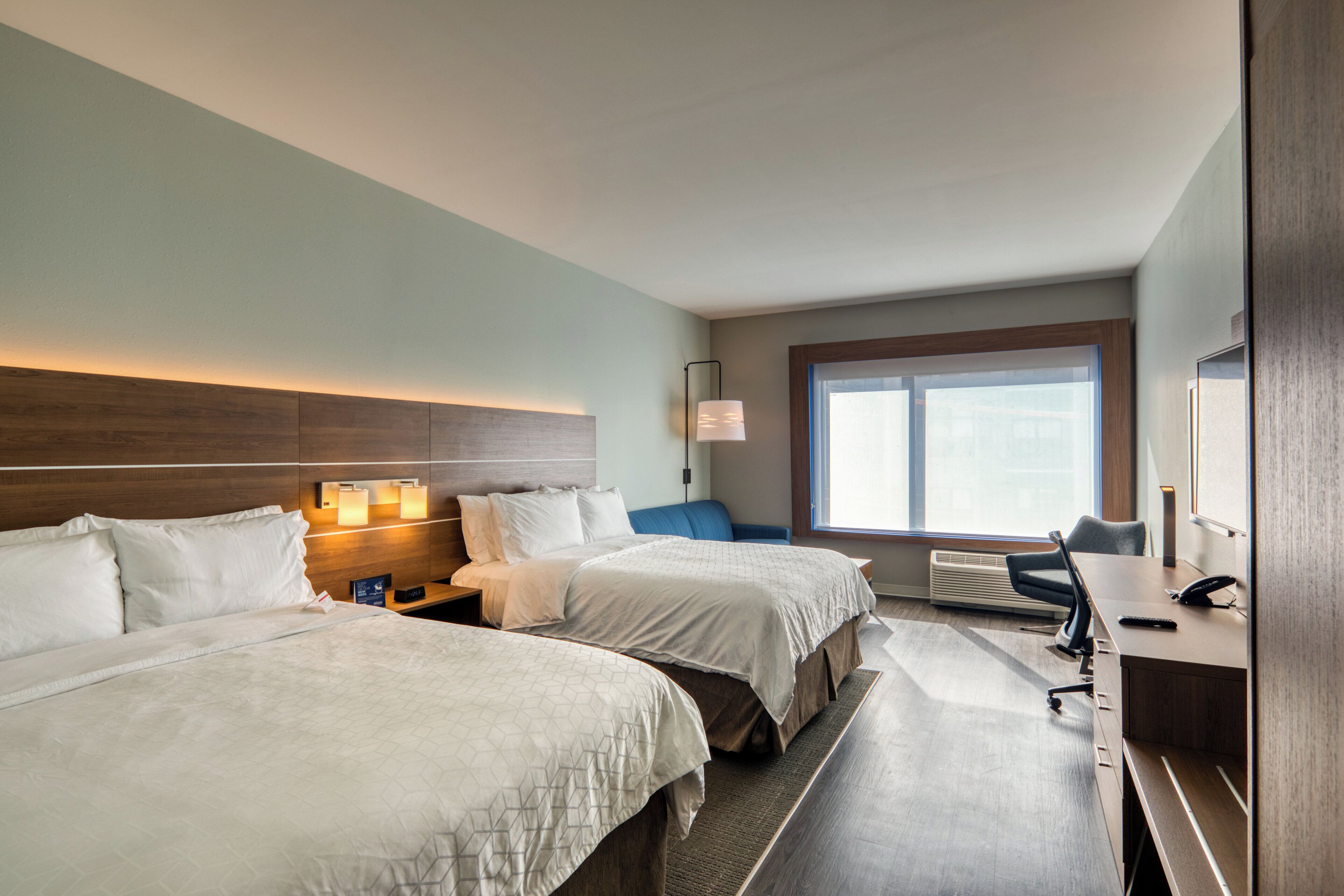 A hotel room with two beds, a desk, a chair, a window, and a nightstand with a lamp.