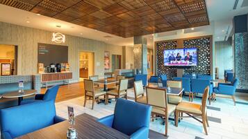 Un moderno vestíbulo de hotel con una barra de café, zonas de estar y una pantalla de televisión mostrando noticias.