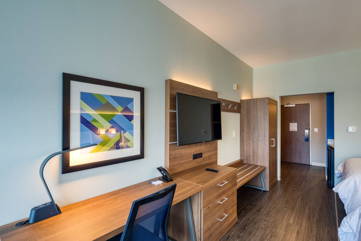 A modern hotel room with a wooden desk, a flat-screen TV, a framed abstract painting, and a bed with white bedding.