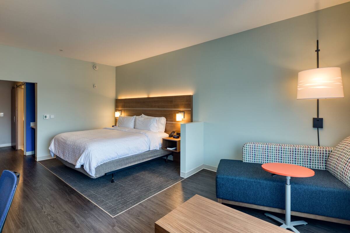A modern hotel room with a bed, bedside table, lamp, and a sofa with a small table.