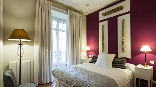 Habitació d'hotel amb un llit gran, dues làmpades laterals, una cadira i vista a la ciutat per la finestra.