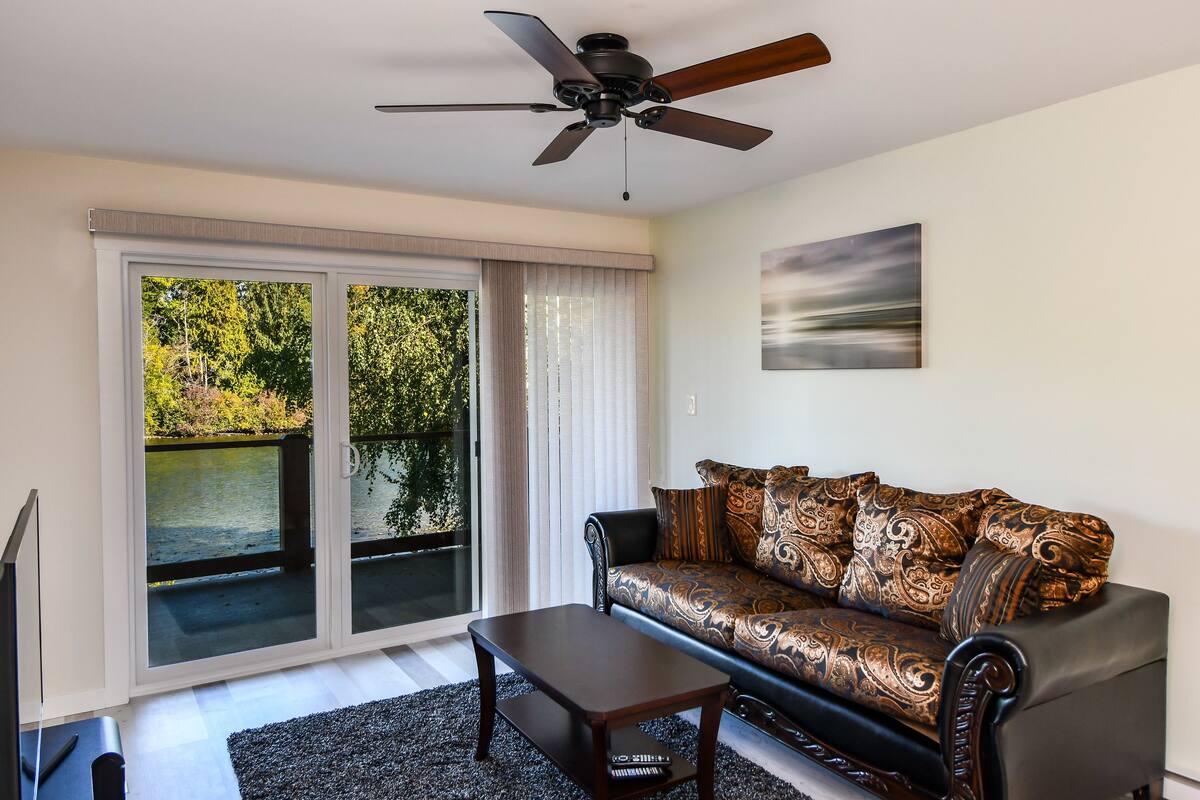 A living room with a sofa, a coffee table, a ceiling fan, and a sliding glass door leading to a balcony with a view of trees and water.