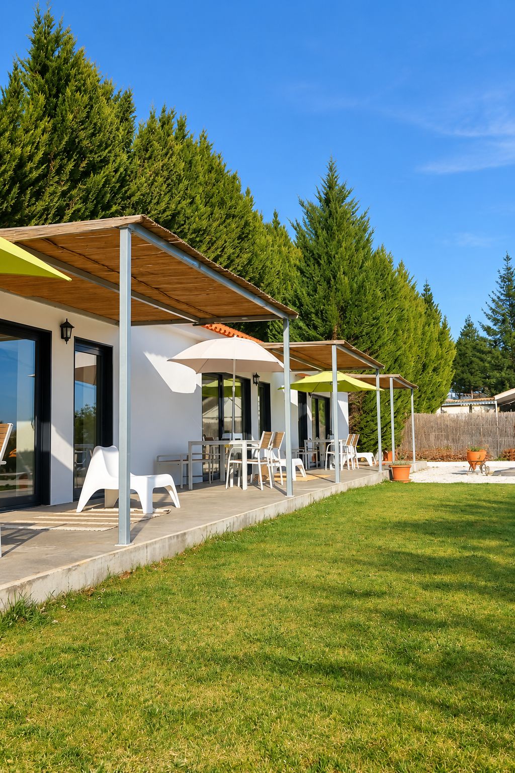 Un área de estar moderna al aire libre con sillas y mesas blancas bajo una pérgola de madera, rodeada de un césped cuidado y árboles altos.