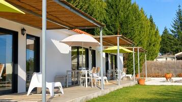 Un área de estar moderna al aire libre con sillas y mesas blancas bajo una pérgola de madera, rodeada de un césped cuidado y árboles altos.