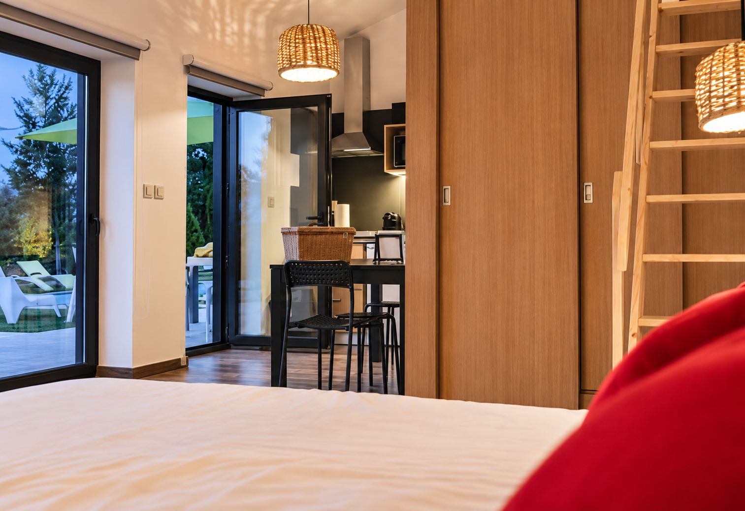 A modern bedroom with a large bed, a dining area with a table and chairs, and a view through a glass door to an outdoor area with lounge chairs.
