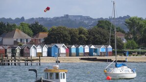A coastal scene with a row of colorful beach huts, a small boat named 'PE-2' anchored near the shore, and a sailboat in the background. - Sandpiper - Moments from Baiter Park (Poole)