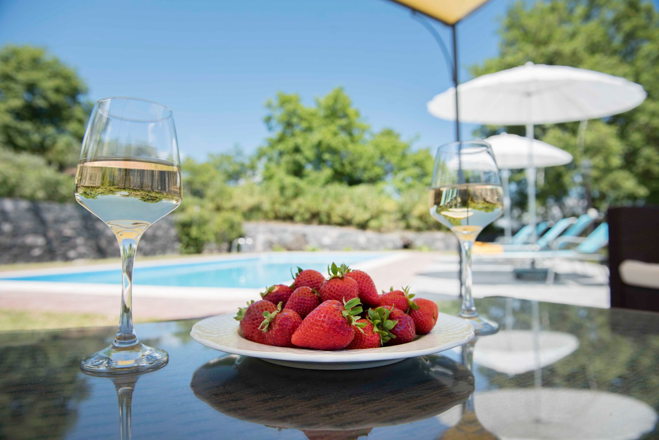 A table with a plate of strawberries and two wine glasses, a pool, and umbrellas in the background.