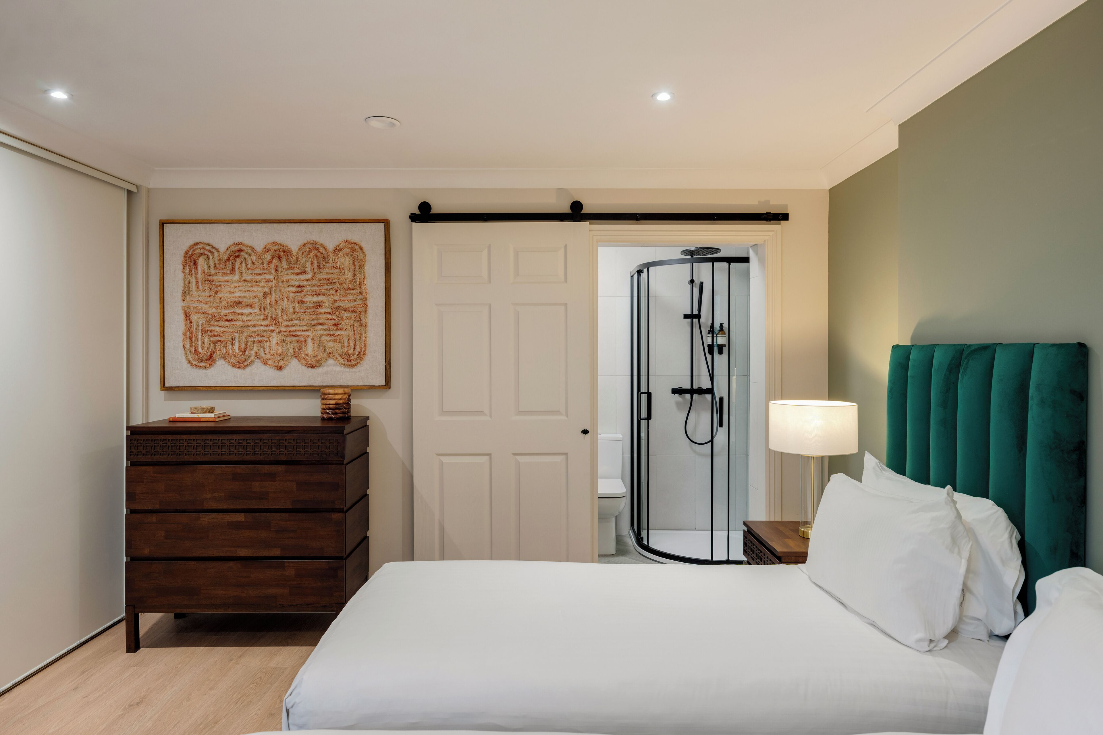 Habitación de hotel con cama, cómoda de madera, obra de arte enmarco, puerta y zona de ducha.