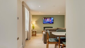 En moderne stue med et fladskærms-TV på væggen, en sofa, et spisebord med stole og et lille sofabord med en lampe.