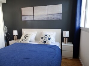 A bedroom with a bed, white nightstands, a wall-mounted fan, and artwork above the bed.