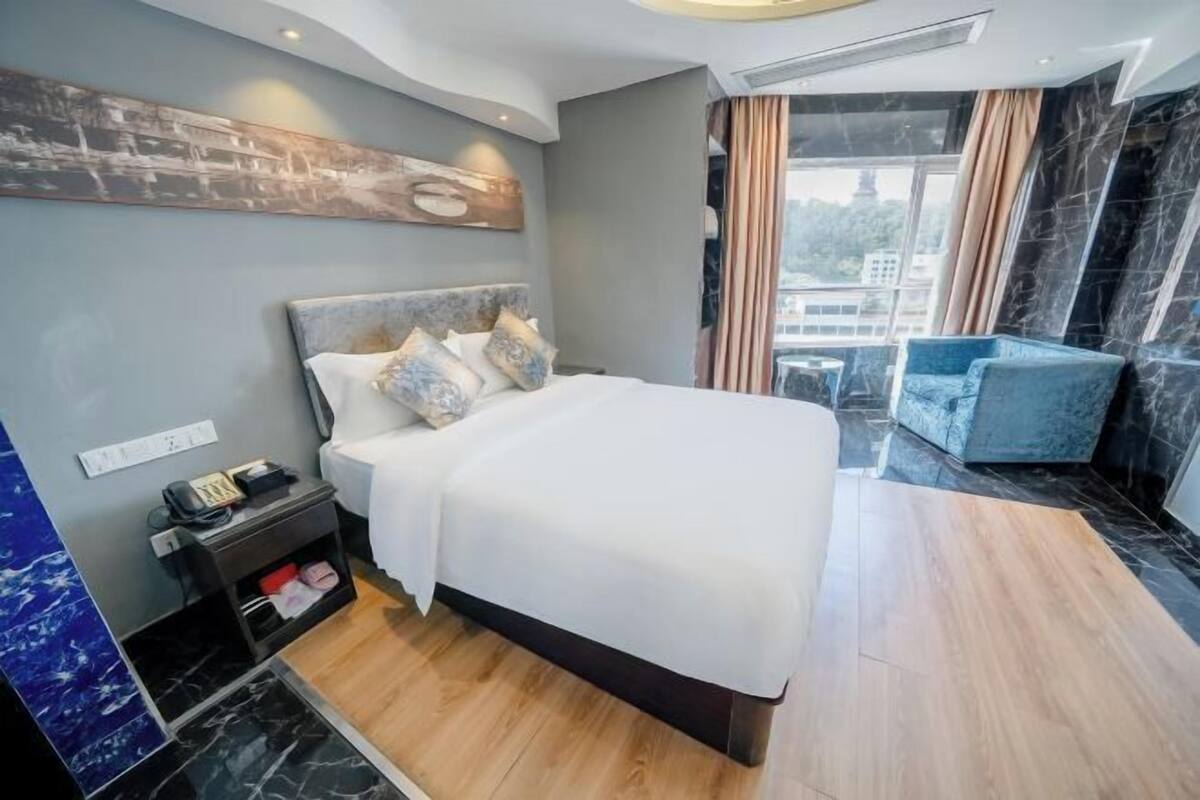 A modern hotel room with a large bed, a bedside table with a phone, a small table, and a view of the city through large windows.