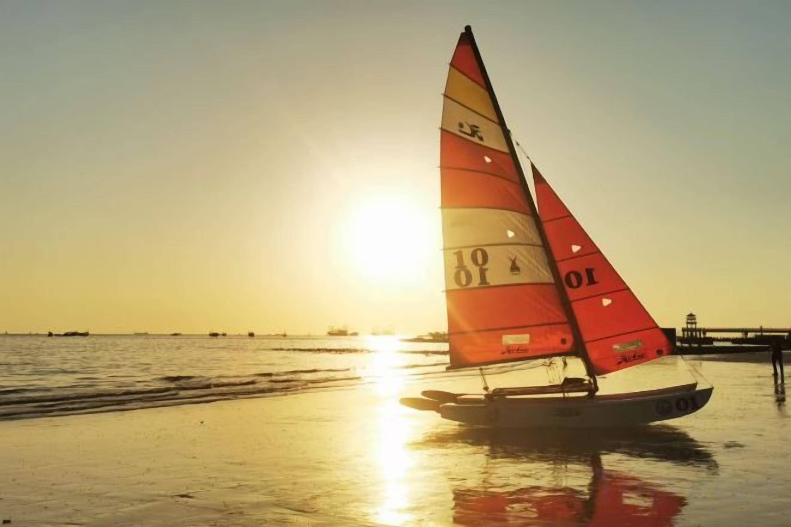 A sailboat with a red sail on a beach during sunset.