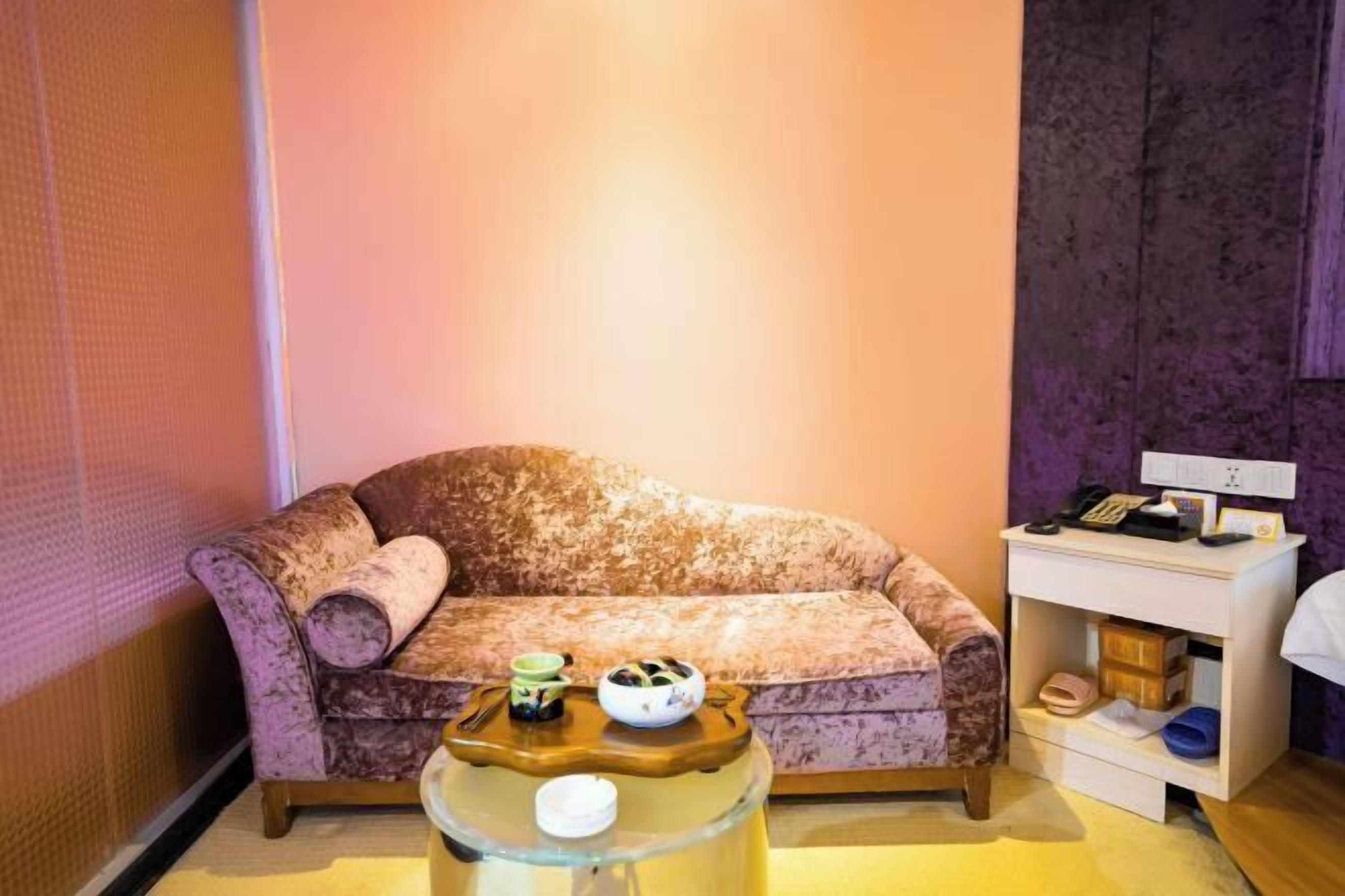 A room with a patterned sofa, a small side table with a bowl and a cup, and a white nightstand with a phone and a lamp.
