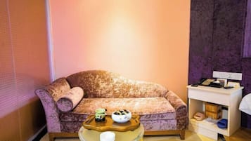 A room with a patterned sofa, a small side table with a bowl and a cup, and a white nightstand with a phone and a lamp.