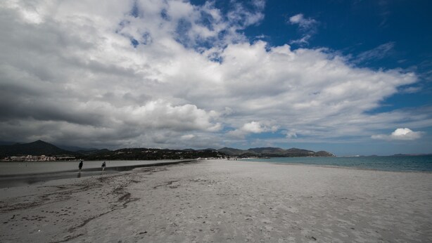 Une vaste plage de sable où quelques personnes se promènent, un ciel dégagé parsemé de nuages et des montagnes lointaines.