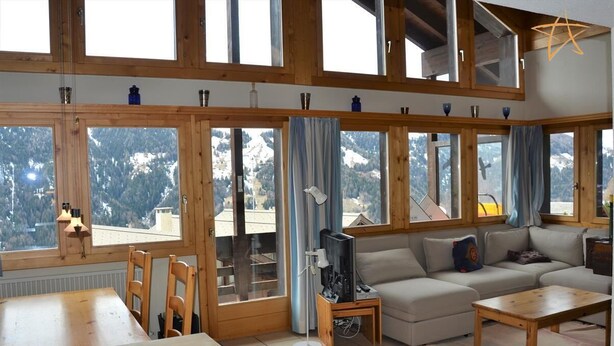 Un accogliente soggiorno con un divano, un tavolino in legno e una televisione. Ampie finestre offrono vista su montagne innevate.