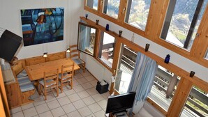 Un interno in legno con un tavolo da pranzo, sedie, televisore e ampie finestre che offrono vista sulle montagne.