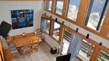Un intérieur en bois comprenant une table à manger, des chaises, un téléviseur et de grandes fenêtres donnant sur une vue imprenable sur les montagnes.