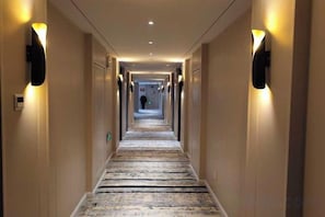Un couloir d’hôtel avec un sol recouvert de moquette, des luminaires fixés au mur et des portes donnant accès aux chambres.
