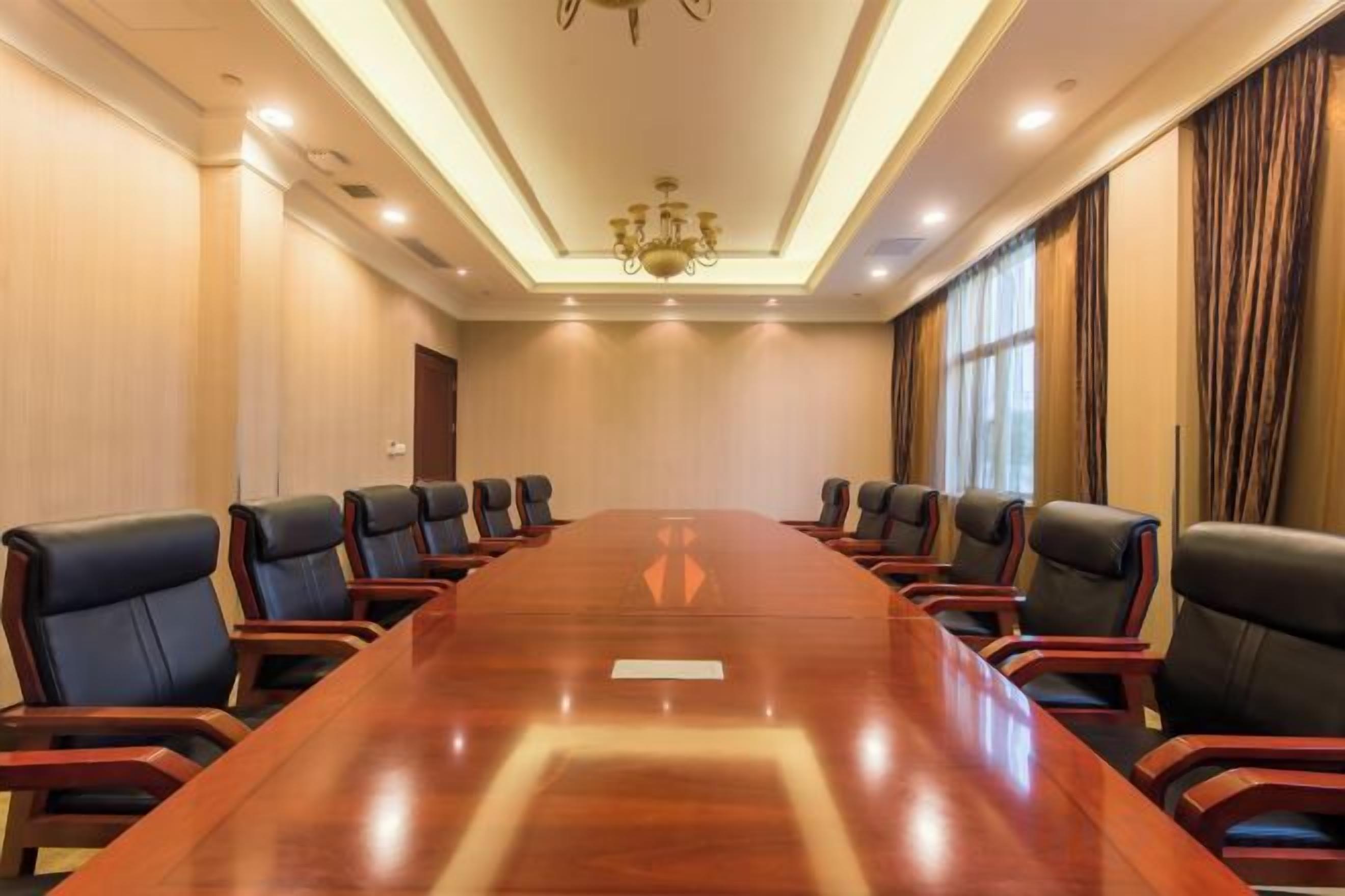 Un salón de conferencias con una larga mesa y múltiples sillas, una araña y ventanas grandes con cortinas.