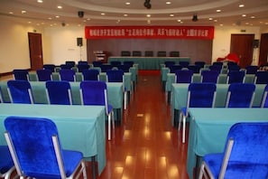 Une salle de conférence avec des rangées de tables et de chaises, une scène avec un pupitre et une banderole accrochée au mur.