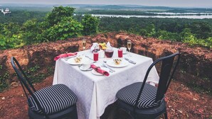 An outdoor dining table set with plates, cutlery, and drinks, overlooking a lush landscape and distant water body.