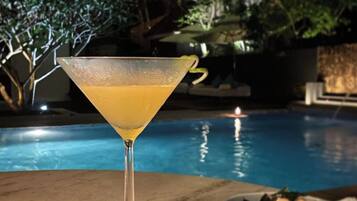 Um copo de martini com uma bebida amarela sobre uma mesa perto de uma piscina, com uma travessa de comida e um apoio decorativo.