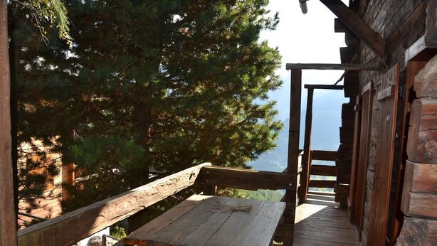 Eine hölzerne Bank und ein Tisch auf einer Holzterrasse mit Blick auf Bäume und einen Berg im Hintergrund.