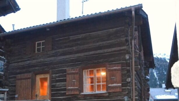 A rustic log cabin with a wooden staircase leading to a front door, a small porch area, and a stone wall with a wooden fence.