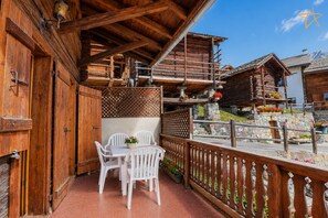Un balcon en bois, agrémenté de chaises et d’une table en plastique, toutes deux de couleur blanche, et offrant une vue sur une région montagneuse parsemée de bâtiments en bois.