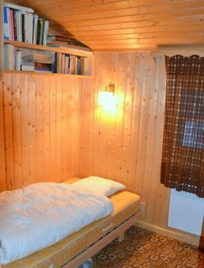 Quarto de madeira com cama, prateleira de livros e luminária na parede.