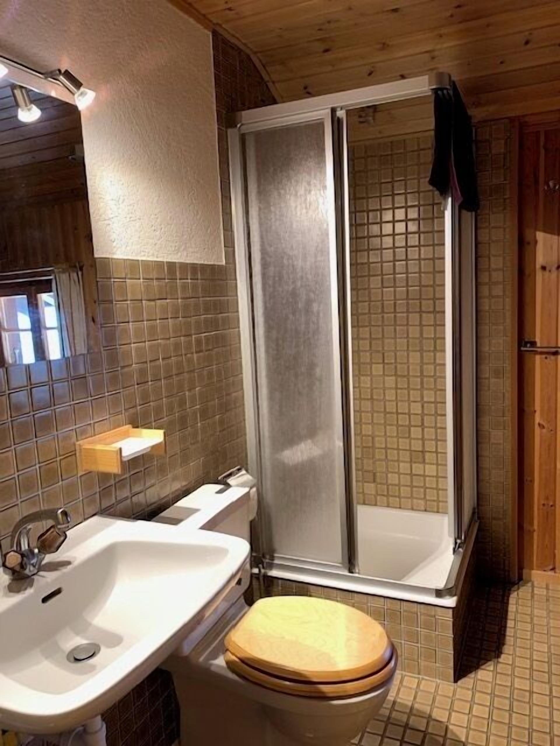 Un bagno con box doccia in legno, un lavandino bianco e uno sgabello del water in legno.