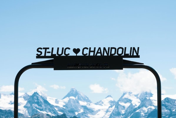 A metal sign with 'ST-LUC CHANDOLIN' and a heart symbol, set against a mountain backdrop.