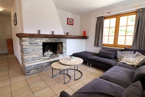 Una sala de estar con chimenea de piedra, un sofá de color oscuro, una mesa redonda de madera y una ventana con cortinas.