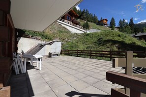 Una terraza con cubierta de madera, mobiliario exterior y vista a una ladera con edificios.