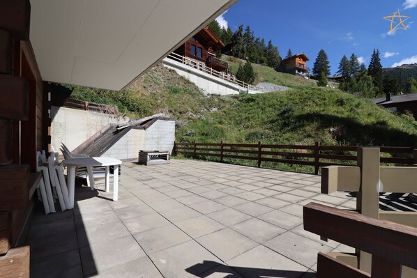 Una terraza con cubierta de madera, mobiliario exterior y vista a una ladera con edificios.