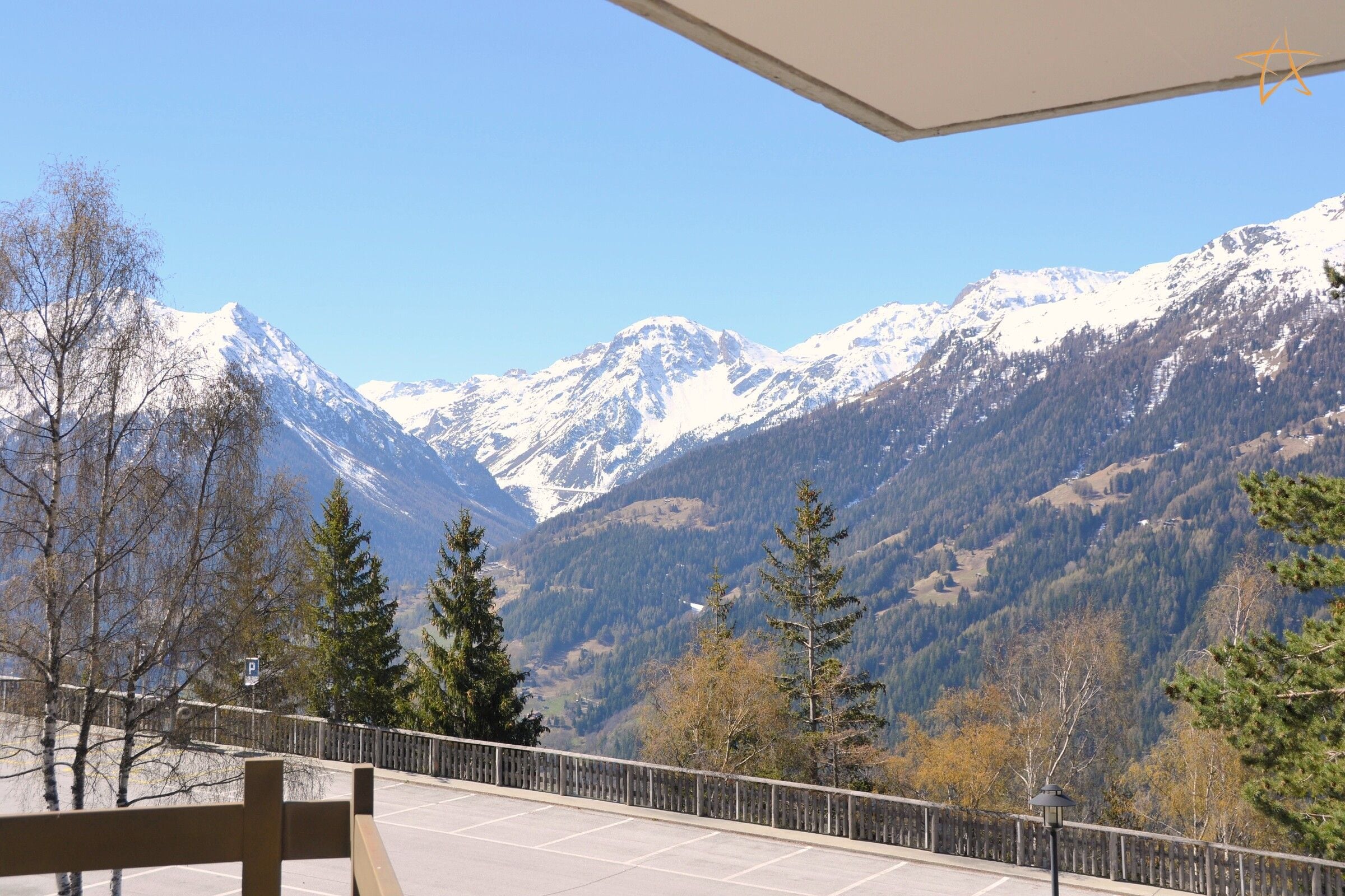 A mountain view from a balcony with a wooden railing, a clear sky, and snow-capped peaks in the distance.