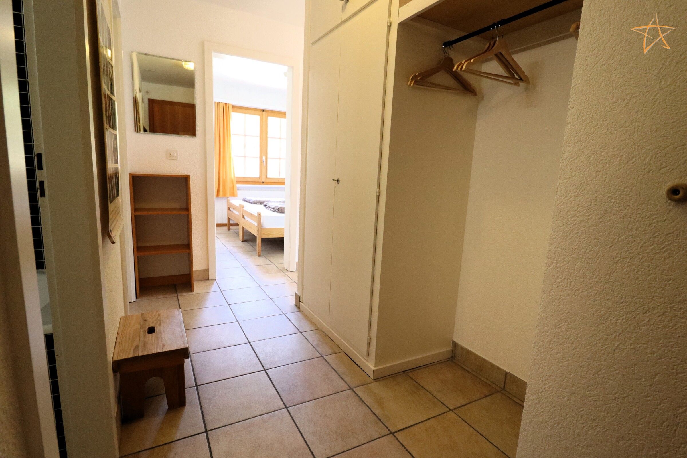 Ein schmaler Flur mit einer hölzernen Sitzbank, einem Kleiderschrank und einem Bett mit einem Fenster im Hintergrund.