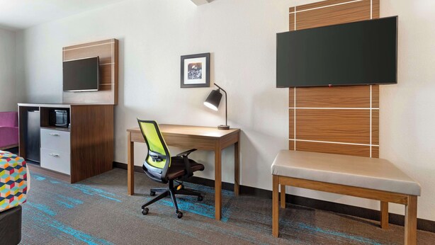 A hotel room with a wooden desk, a chair, a TV mounted on the wall, a framed picture, and a bed with a colorful geometric pillow. - Best Western Plus Medical Center (Amarillo)
