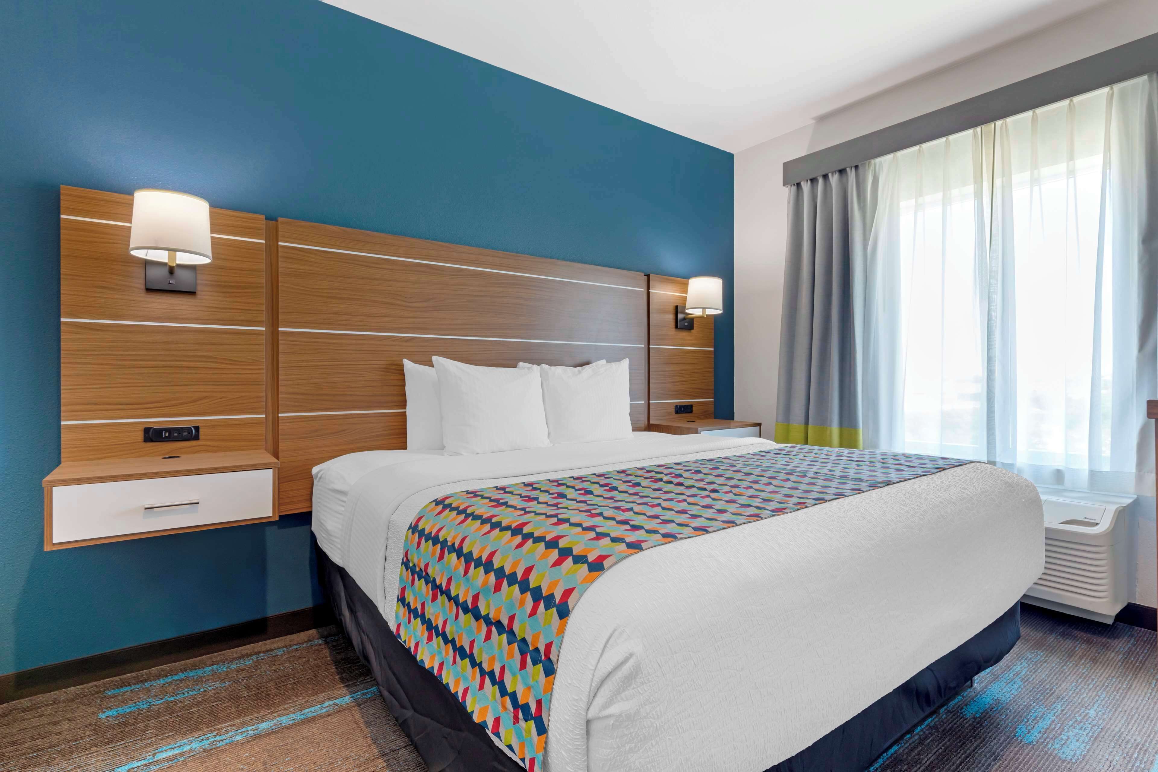 A hotel room with a large bed, a wooden headboard, two bedside tables with lamps, a window with curtains, and a blue accent wall.